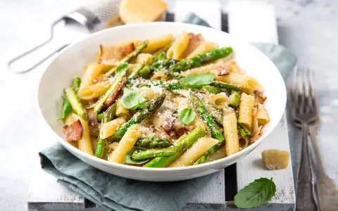 Piatto di pasta con asparagi e pancetta croccante, guarnito con basilico fresco e parmigiano grattugiato.