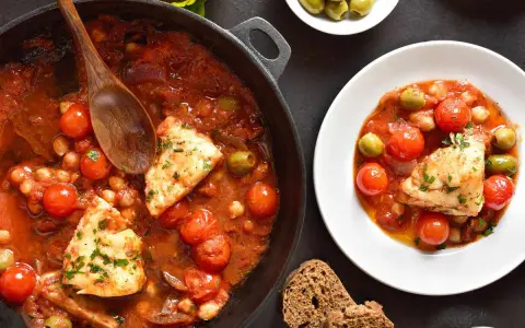 Merluzzo in umido con pomodorini, olive e ceci, servito in una padella e in un piatto, con pane scuro.