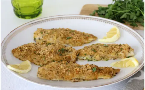 Four breaded fish fillets on a white plate with lemon wedges and parsley.