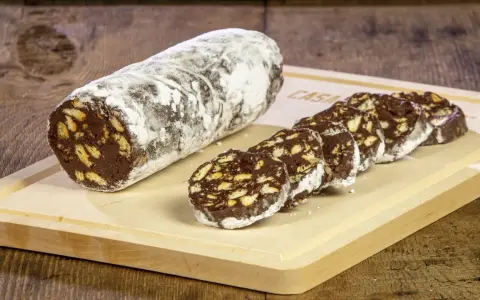 Un rotolo di salame al cioccolato, cosparso di zucchero a velo, affiancato da fette tagliate su un tagliere di legno.