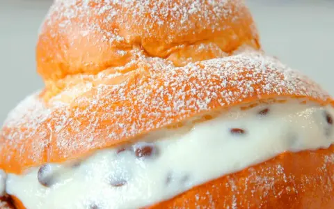 Brioche col tuppo soffice e dorata, ripiena di crema vellutata con gocce di cioccolato, spolverata di zucchero a velo.