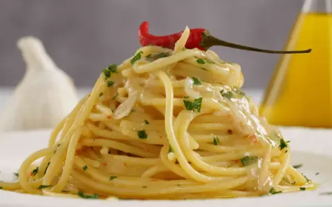 Spaghetti conditi con aglio, olio e peperoncino, guarniti con prezzemolo e un peperoncino rosso. Sullo sfondo, un bulbo d'aglio e una bottiglia d'olio.