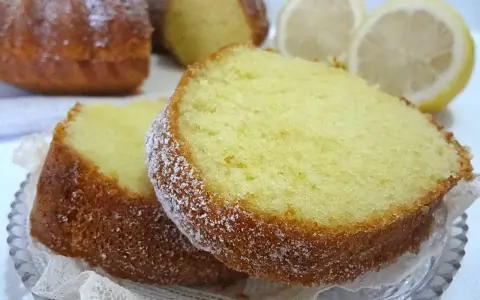 Fette di un soffice ciambellone al limone, spolverate di zucchero a velo, con spicchi di limone sullo sfondo.