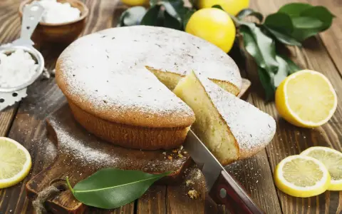 Torta al limone velocissima, spolverata di zucchero a velo, con una fetta tagliata e limoni freschi intorno.