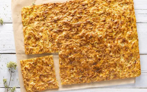 Schiacciata di zucchine dorata e fragrante, tagliata a pezzi su carta da forno, con erbe aromatiche sparse.