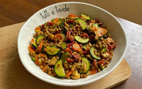 Insalata di lenticchie colorata con zucchine, carote e pomodori secchi, servita in una ciotola con la scritta "tutti a tavola".