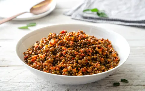 Un piatto invitante di lenticchie in umido, condite con pomodoro e cipolla, servite in una ciotola bianca su un tavolo di legno.