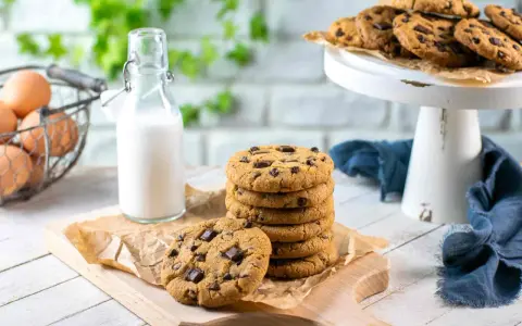 Una pila di biscotti con gocce di cioccolato su un tagliere, accanto a una bottiglia di latte e un cestino di uova.