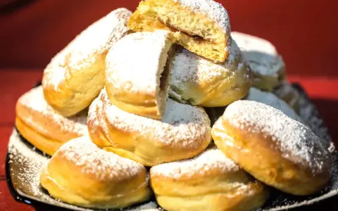 Pile di bomboloni ricetta originale, soffici e cosparsi di zucchero a velo. Uno è tagliato a metà, rivelando un ripieno goloso.
