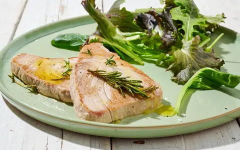 Due fette di tonno in padella, condite con olio e rosmarino, servite con insalata fresca su un piatto verde.