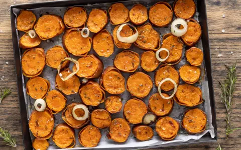 Fette di patate dolci al forno, dorate e condite con erbe aromatiche e sale, disposte su una teglia con anelli di cipolla.