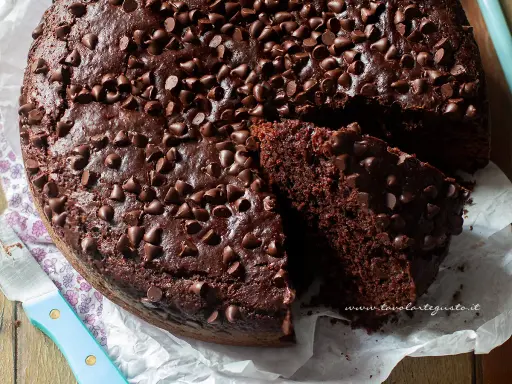 A rich, moist chocolate cake topped with chocolate chips, with a slice cut out to show its texture.