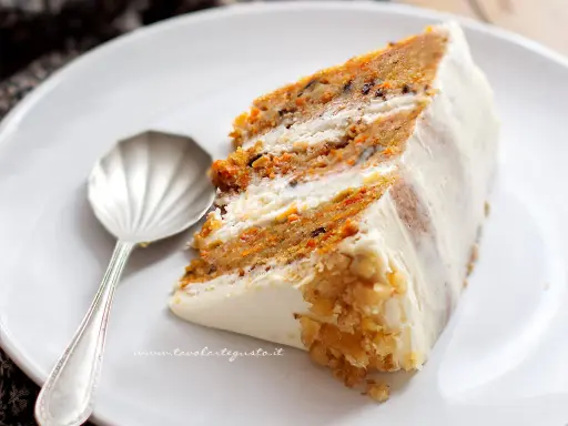 Una fetta di torta di carote e yogurt, soffice e profumata, con glassa bianca e noci tritate.