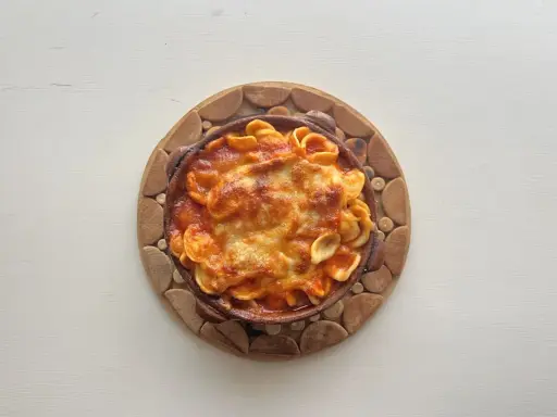 Orecchiette al forno in terracotta, con sugo rosso e formaggio filante, servite su un tagliere di legno.