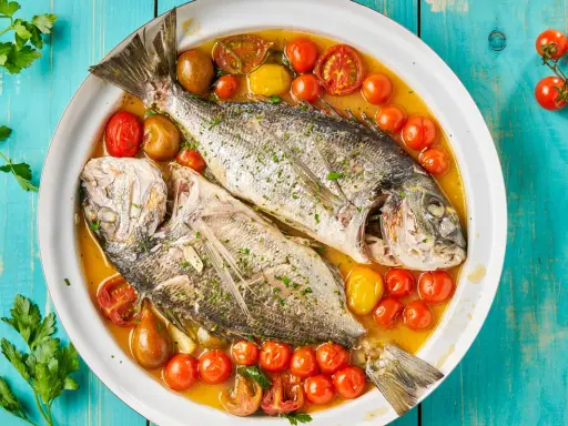 Orata all'acqua pazza, due pesci interi cotti in un brodo saporito con pomodorini e erbe aromatiche, serviti in un piatto bianco su un tavolo di legno blu.