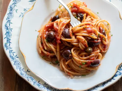 Un piatto di spaghetti alla puttanesca, con pomodoro, olive nere e capperi, servito su un elegante piatto bianco con decorazioni blu.