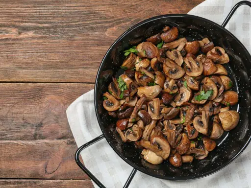 A black pan filled with perfectly cooked, sliced mushrooms and garnished with parsley, sits on a wooden table.