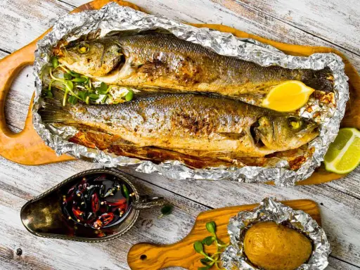 Due branzini cotti al cartoccio, dorati e profumati, serviti con limone, patata al cartoccio e salsa piccante.