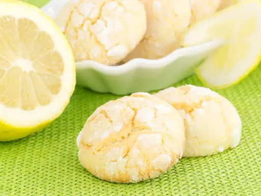 Biscotti al limone spolverati di zucchero a velo, accanto a fette di limone fresco su sfondo verde.