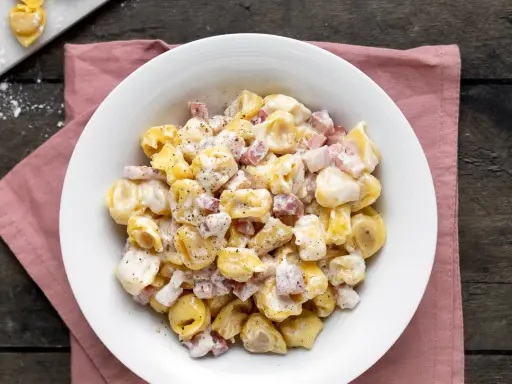Un piatto invitante di tortellini panna e prosciutto, conditi con pepe nero, su un tovagliolo rosa.