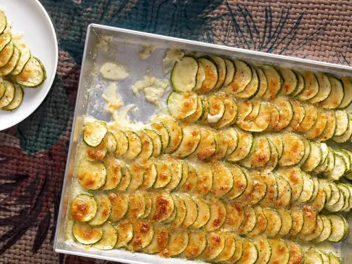 Fette di zucchine al forno con mozzarella filante, disposte in file ordinate in una teglia e su un piatto bianco.