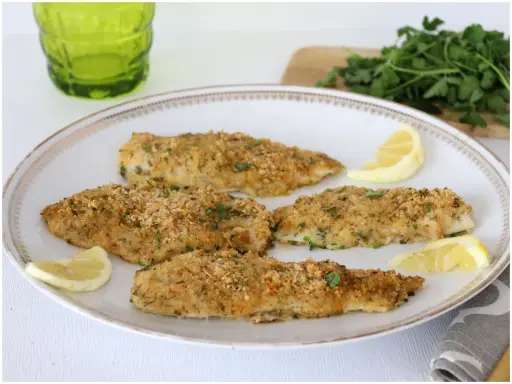 Four breaded fish fillets on a white plate with lemon wedges and parsley.