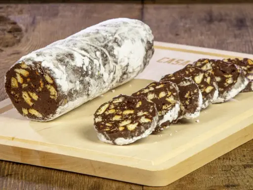 Un rotolo di salame al cioccolato, cosparso di zucchero a velo, affiancato da fette tagliate su un tagliere di legno.