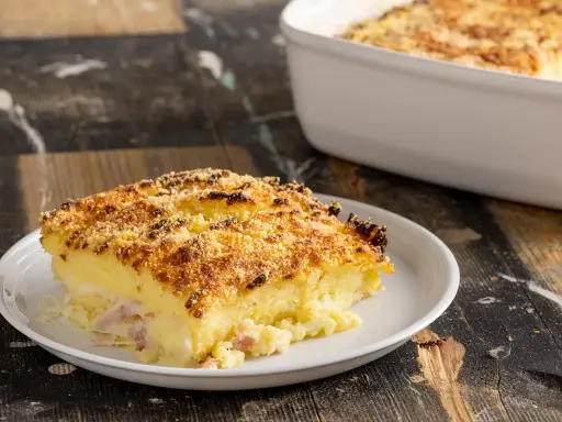 Una fetta di sformato di patate dorato e croccante, con prosciutto e formaggio filante, servita su un piatto bianco.