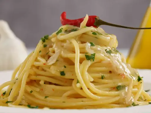 Spaghetti conditi con aglio, olio e peperoncino, guarniti con prezzemolo e un peperoncino rosso. Sullo sfondo, un bulbo d'aglio e una bottiglia d'olio.