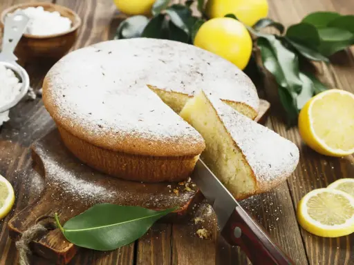 Torta al limone velocissima, spolverata di zucchero a velo, con una fetta tagliata e limoni freschi intorno.