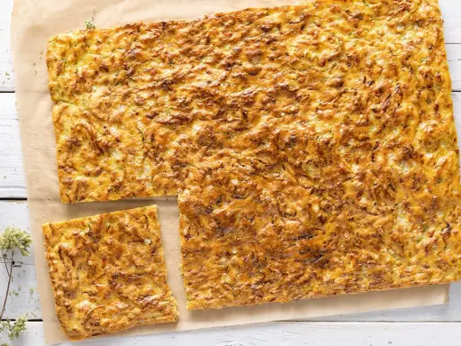 Schiacciata di zucchine dorata e fragrante, tagliata a pezzi su carta da forno, con erbe aromatiche sparse.