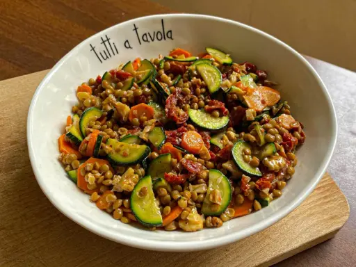 Insalata di lenticchie colorata con zucchine, carote e pomodori secchi, servita in una ciotola con la scritta "tutti a tavola".