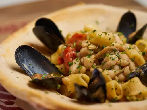 Un piatto invitante di pasta fagioli e cozze, con pomodorini e prezzemolo fresco, servito in una ciotola di pane croccante.