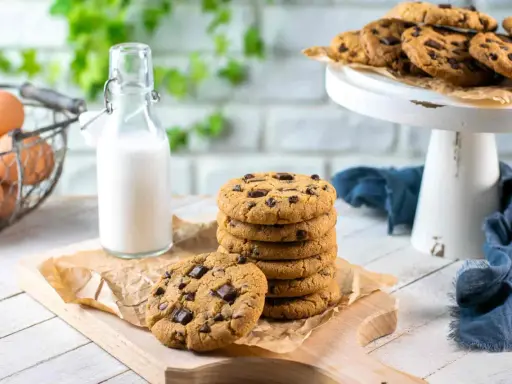 Una pila di biscotti con gocce di cioccolato su un tagliere, accanto a una bottiglia di latte e un cestino di uova.