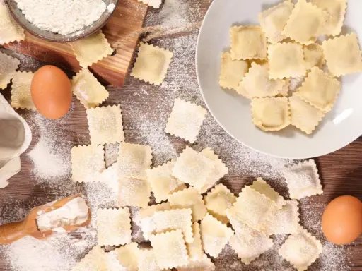 Tanti ravioli fatti in casa sparsi su un tavolo di legno, con farina, uova e una paletta.