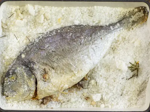 Orata al sale, intera e dorata, adagiata su un letto di sale grosso e rosmarino in una teglia rossa.