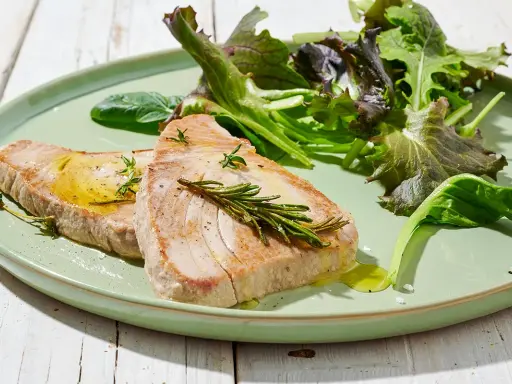 Due fette di tonno in padella, condite con olio e rosmarino, servite con insalata fresca su un piatto verde.