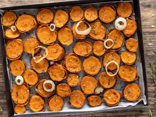 Fette di patate dolci al forno, dorate e condite con erbe aromatiche e sale, disposte su una teglia con anelli di cipolla.