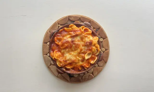 Orecchiette al forno in terracotta, con sugo rosso e formaggio filante, servite su un tagliere di legno.