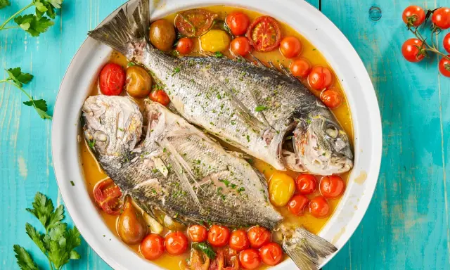 Orata all'acqua pazza, due pesci interi cotti in un brodo saporito con pomodorini e erbe aromatiche, serviti in un piatto bianco su un tavolo di legno blu.