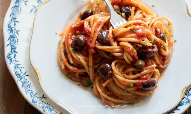 Un piatto di spaghetti alla puttanesca, con pomodoro, olive nere e capperi, servito su un elegante piatto bianco con decorazioni blu.