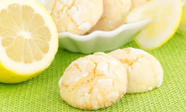 Biscotti al limone spolverati di zucchero a velo, accanto a fette di limone fresco su sfondo verde.