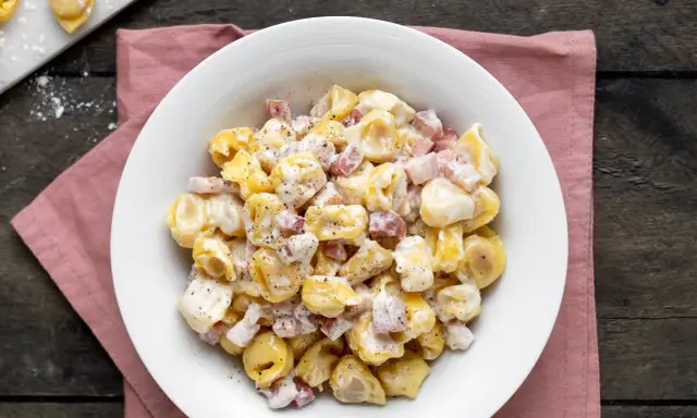 Un piatto invitante di tortellini panna e prosciutto, conditi con pepe nero, su un tovagliolo rosa.