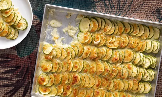 Fette di zucchine al forno con mozzarella filante, disposte in file ordinate in una teglia e su un piatto bianco.