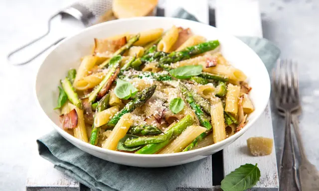 Piatto di pasta con asparagi e pancetta croccante, guarnito con basilico fresco e parmigiano grattugiato.