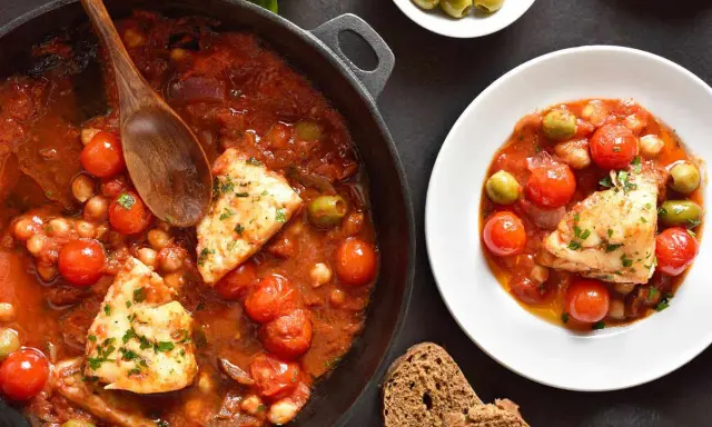 Merluzzo in umido con pomodorini, olive e ceci, servito in una padella e in un piatto, con pane scuro.