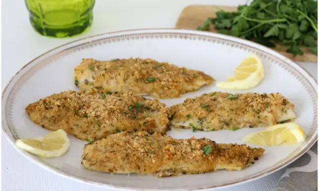 Four breaded fish fillets on a white plate with lemon wedges and parsley.