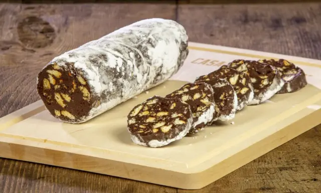 Un rotolo di salame al cioccolato, cosparso di zucchero a velo, affiancato da fette tagliate su un tagliere di legno.