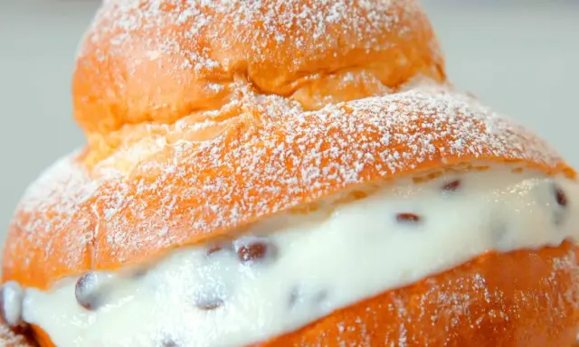 Brioche col tuppo soffice e dorata, ripiena di crema vellutata con gocce di cioccolato, spolverata di zucchero a velo.