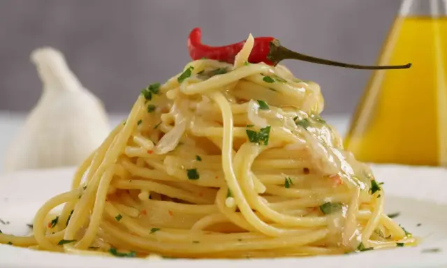 Spaghetti conditi con aglio, olio e peperoncino, guarniti con prezzemolo e un peperoncino rosso. Sullo sfondo, un bulbo d'aglio e una bottiglia d'olio.